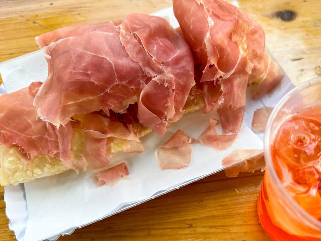 White platter with gnocco fritto topped with prosciutto crudo. Aperol spritz in lower right corner.