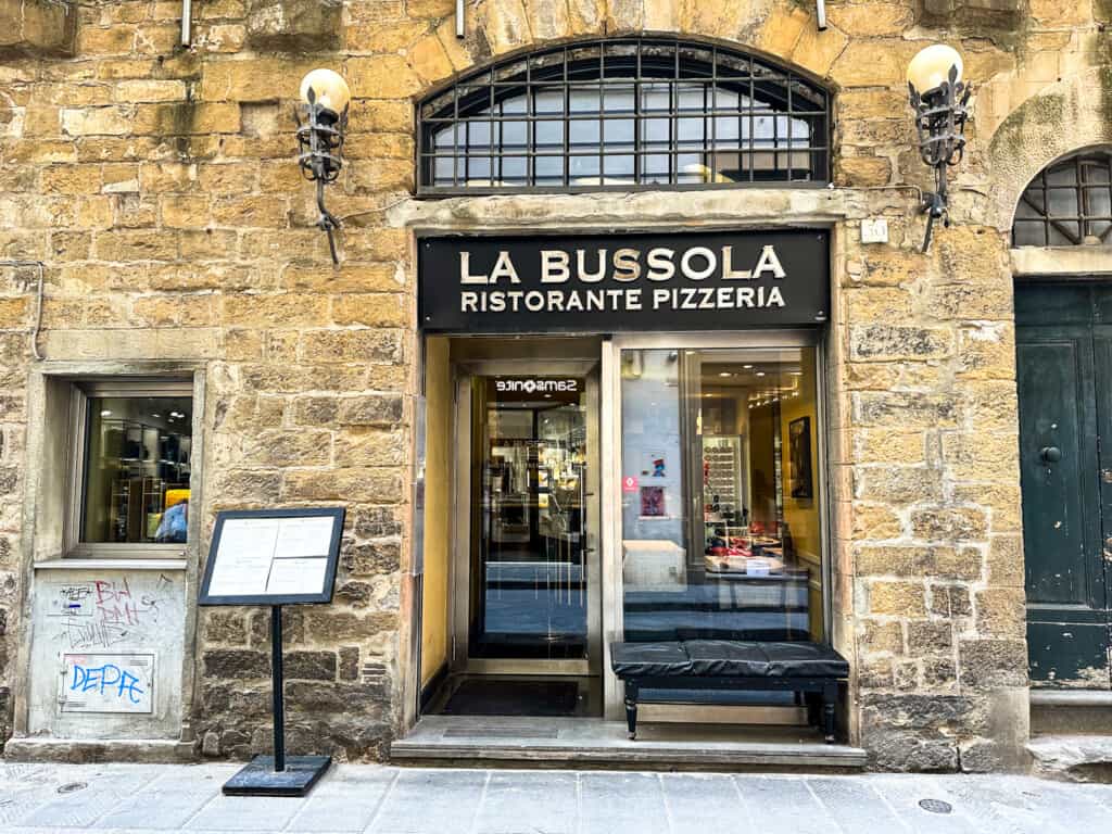 side street view of la bussola pizzeria with sign overhead of entrance and a menu on a stand to the left side of the door surrounded by brick wall