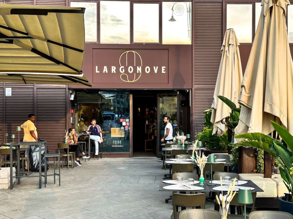 side view of restaurant largo nove with brown sign hanging in center with tables outside set for dinner on either side on a dark grey terrace