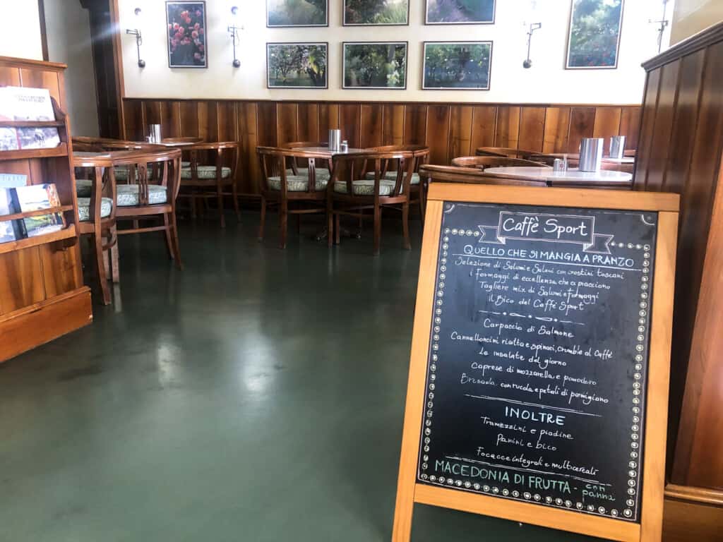 View inside a bar in Italy. Wooden tables and chairs and a sign on right for 'Caffè Sport' with a handwritten menu in Italian.