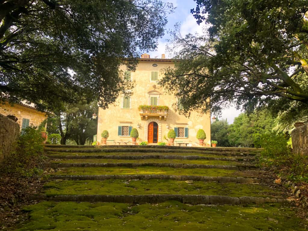 Yellow Italian building at top of moss-covered stone steps. Trees on either side and potted plants decorate the entrance.