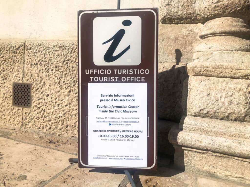 Sign for tourist office that lists hours. Background is stone building.