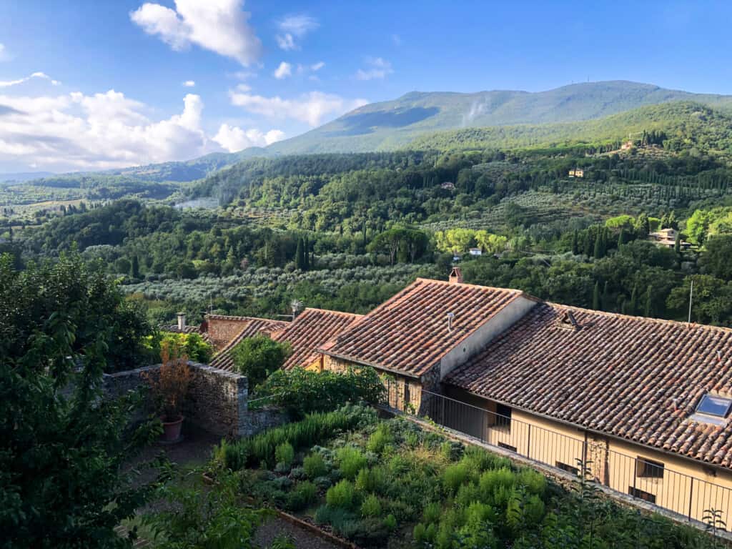 View of lush green countryside in Tuscany. You can see green-covered mountain in background. In foreground is Italian home with roof covered in terracotta tiles.