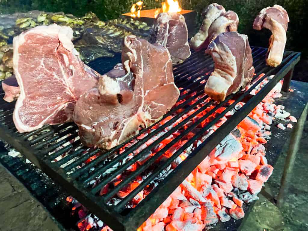 side view of several bistecche sitting on bone straight up over a grill on hot coals outdoors from side view with grass in background.