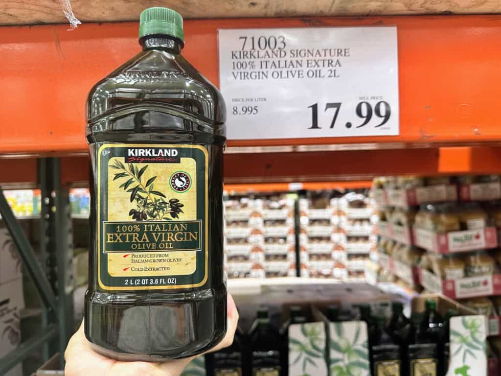 Hand holding large bottle of Costco's Kirkland olive oil next to sign with price.