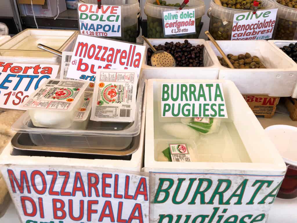 side view of various white containers of different kinds of mozzarella for sale on a table at an outdoor market with different colored signs indicating type.