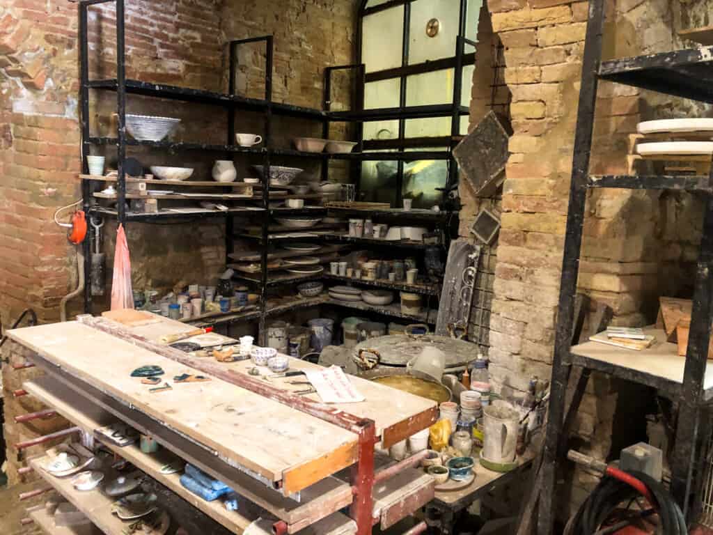 side view of indoor Pippo's ceramic studio from indoors with large table and shelving in background with various pots to paint with colors everywhere. 