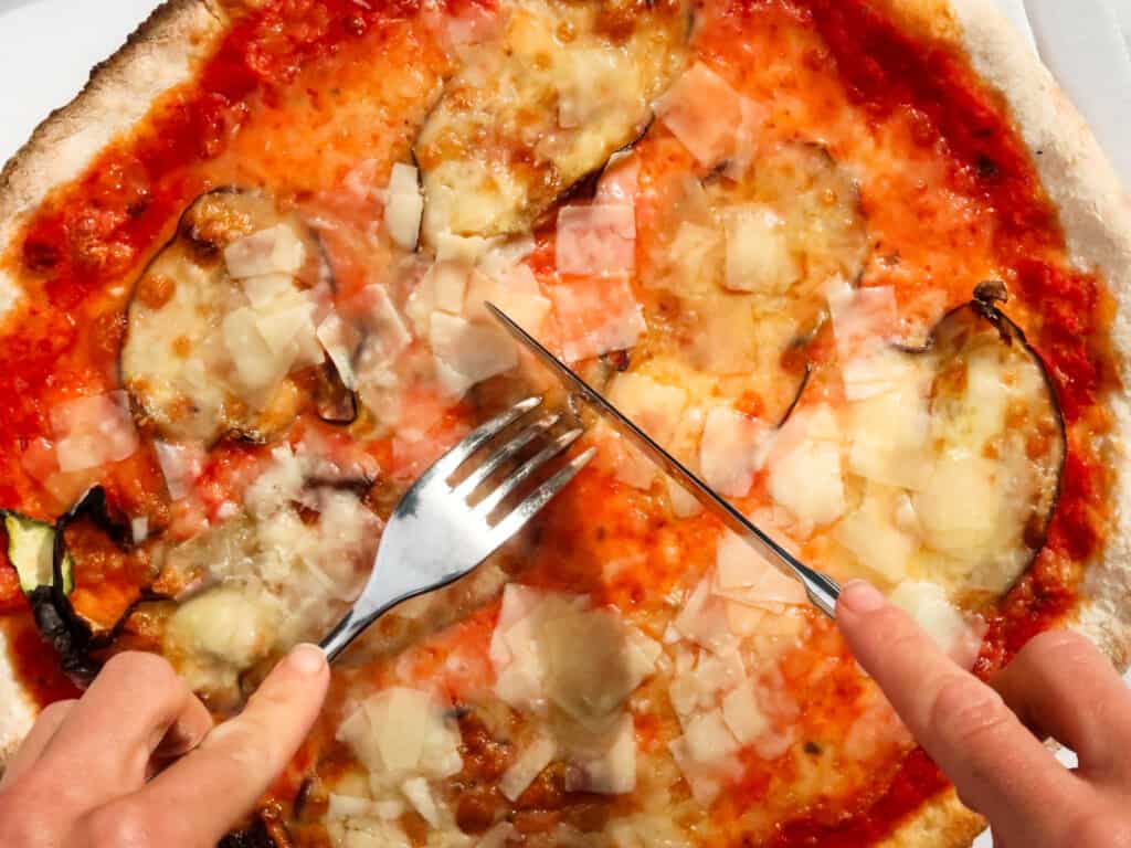 Hands holding fork and knife over a pizza with eggplant and parmigiano toppings.