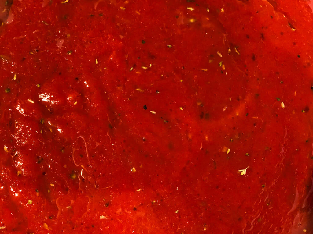 top image up close of bright red pizza sauce with specs of oregano and oil drops