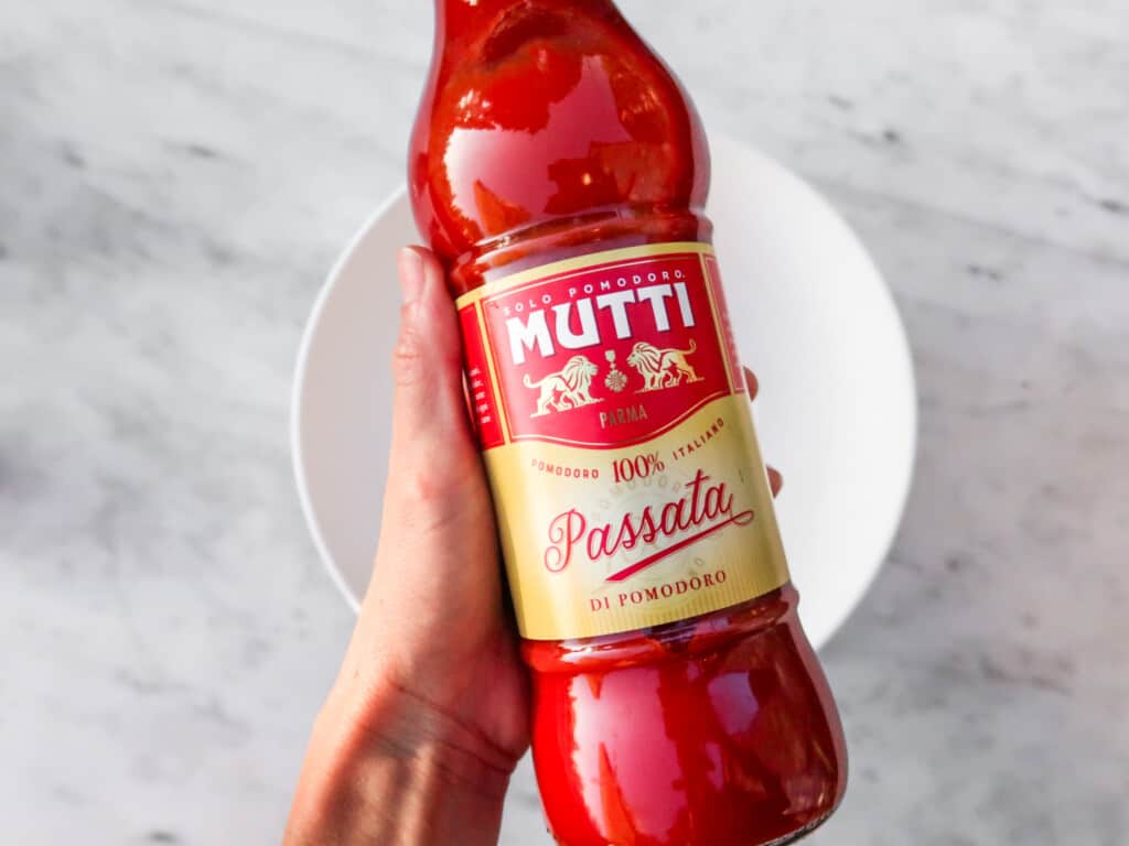 hand holding a jar of mutti passata tomato over a white bowl on a marble background from top view