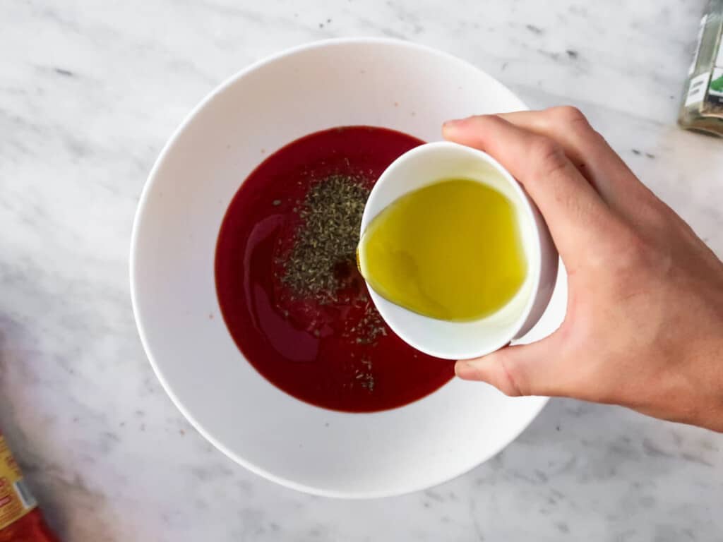 hand holding a small white bowl of olive oil and pouring it into a white bowl of tomato sauce on a marble table from top view