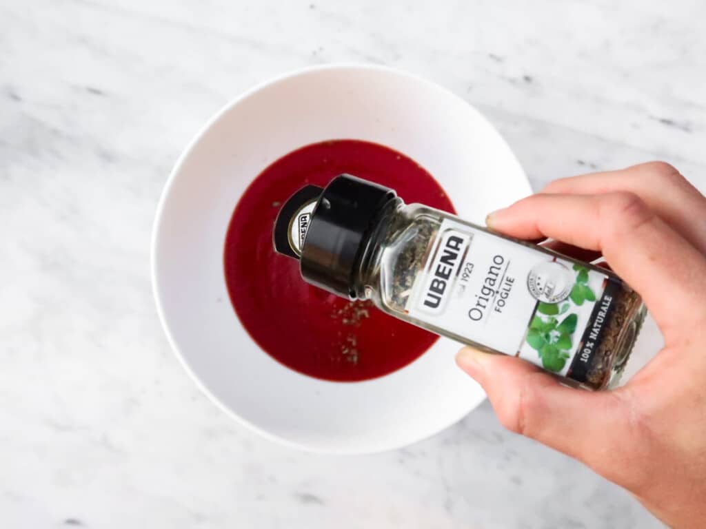 hand holding a small dispenser of oregano and pouring it into a white bowl with tomato sauce on a marble table from top view