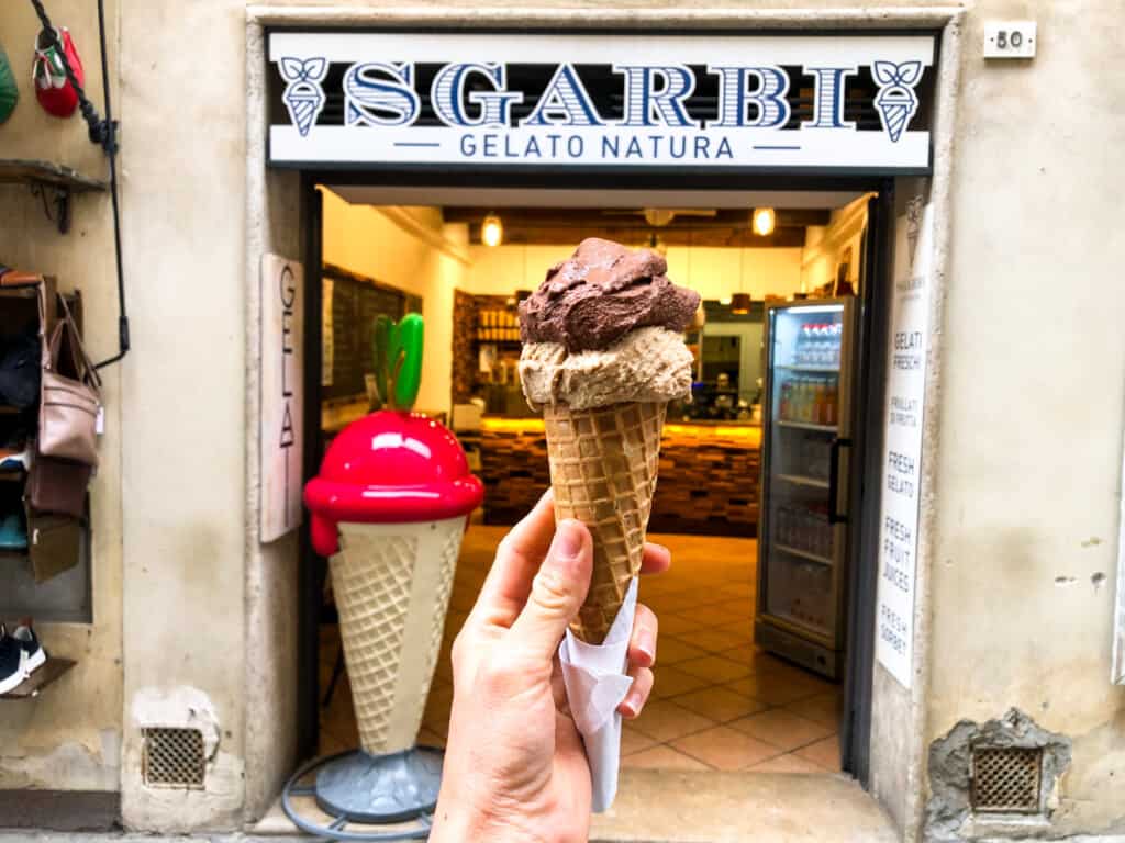 hand holding a cone with chocolate and dark brown coffee ice cream in a cone at arms reach infront of sgarbi sign outside of gelateria on street with beige walls.