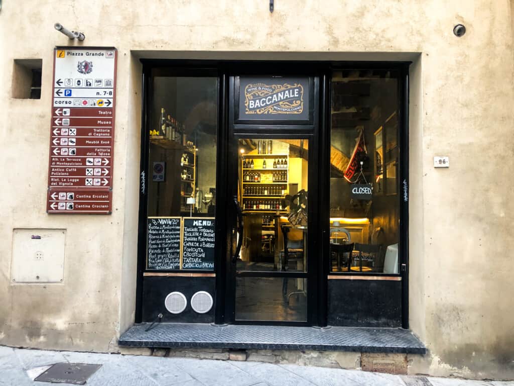 Entrance to Baccanale wine shop in Montepulciano. Glass windows and door with handwritten chalk sign in the window. To left on wall are street and directional signs for the town.