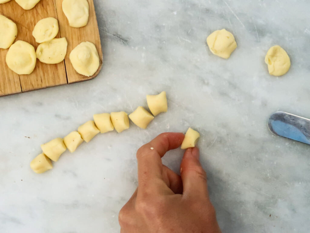 Index finger and thumb hold dough ready to be formed into orecchiette pasta. You can see the finished results on a wooden cutting board and the marble surface.