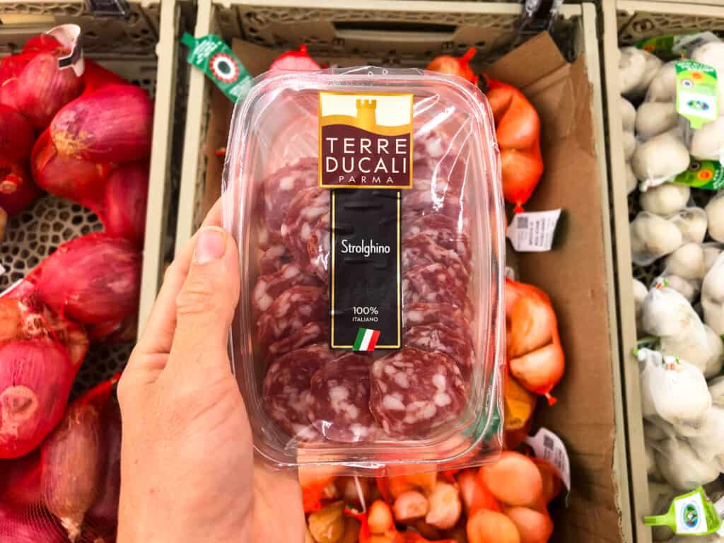 Single portion of salame in a plastic container at the supermarket. A hand holds it above bins of garlic and shallots.