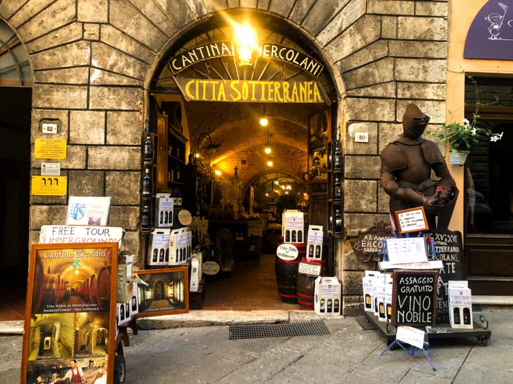 Entrance to Cantina Ercolani in Montepulciano, Italy. There is knight's armor to the right of the entrance. Wine bottles decorate the shelves indoors and are displayed in boxes outside the entrance.