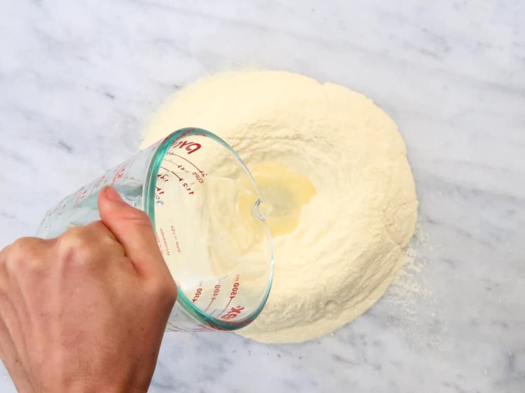 Hand using glass measuring cup to pour water into flour to make pasta dough.