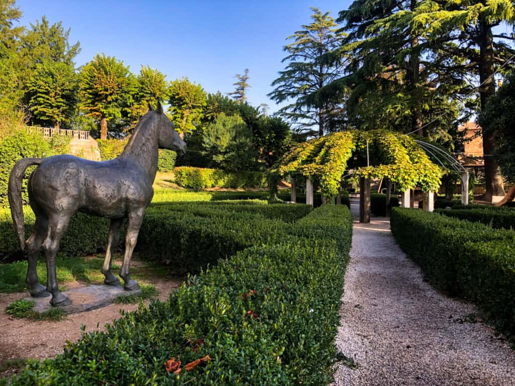 Metal statue of horse in garden area in Montepulciano. There are low hedges and trees.