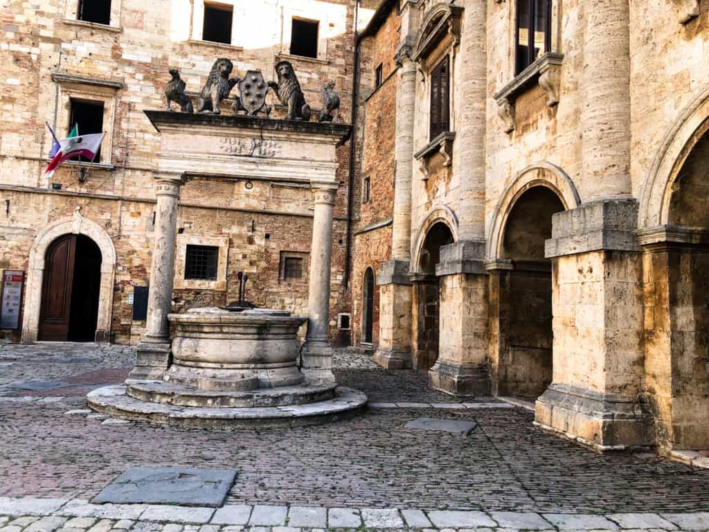The main fountain with lion statues on top and sand-colored stone buildings in Piazza Grande in Montepulciano, Italy.