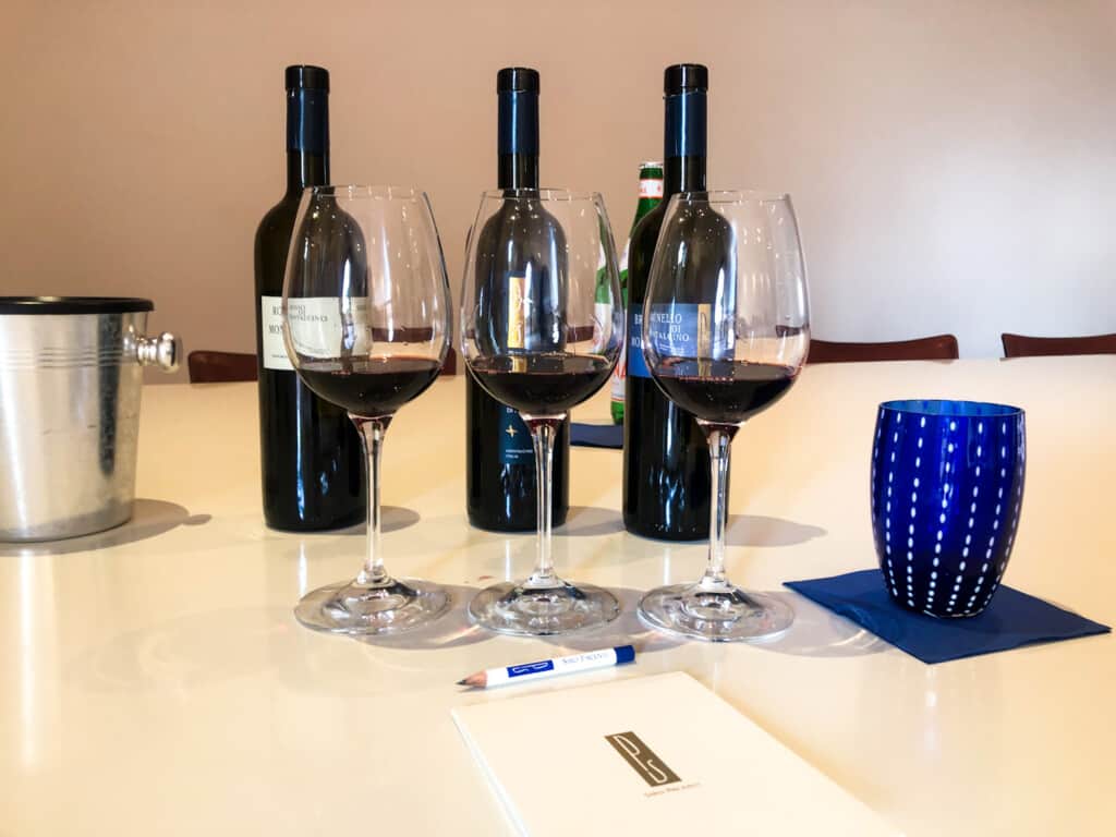 Three wine glasses sit in front of three wine bottles on a white table. Blue glass on napkin to the right and pencil and paper in front.