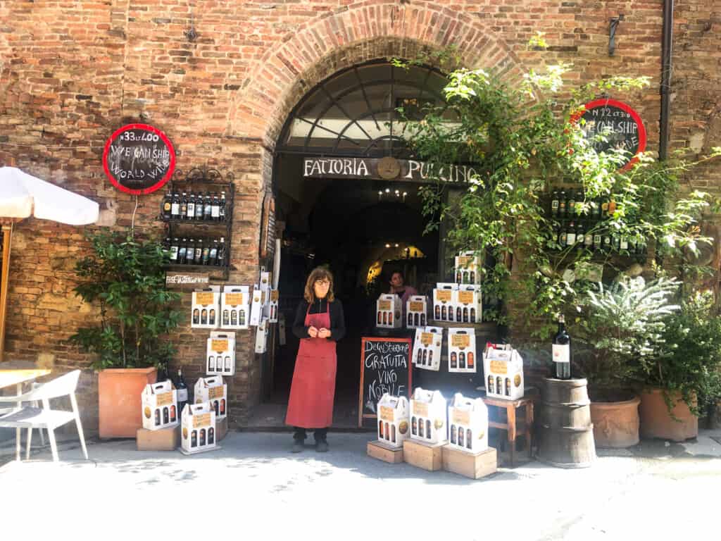 Entrance to an enoteca in Montepulciano, Italy. Woman in red apron standing in front of arched entrance. Plants and wine bottles decorate the area in front of the shop.