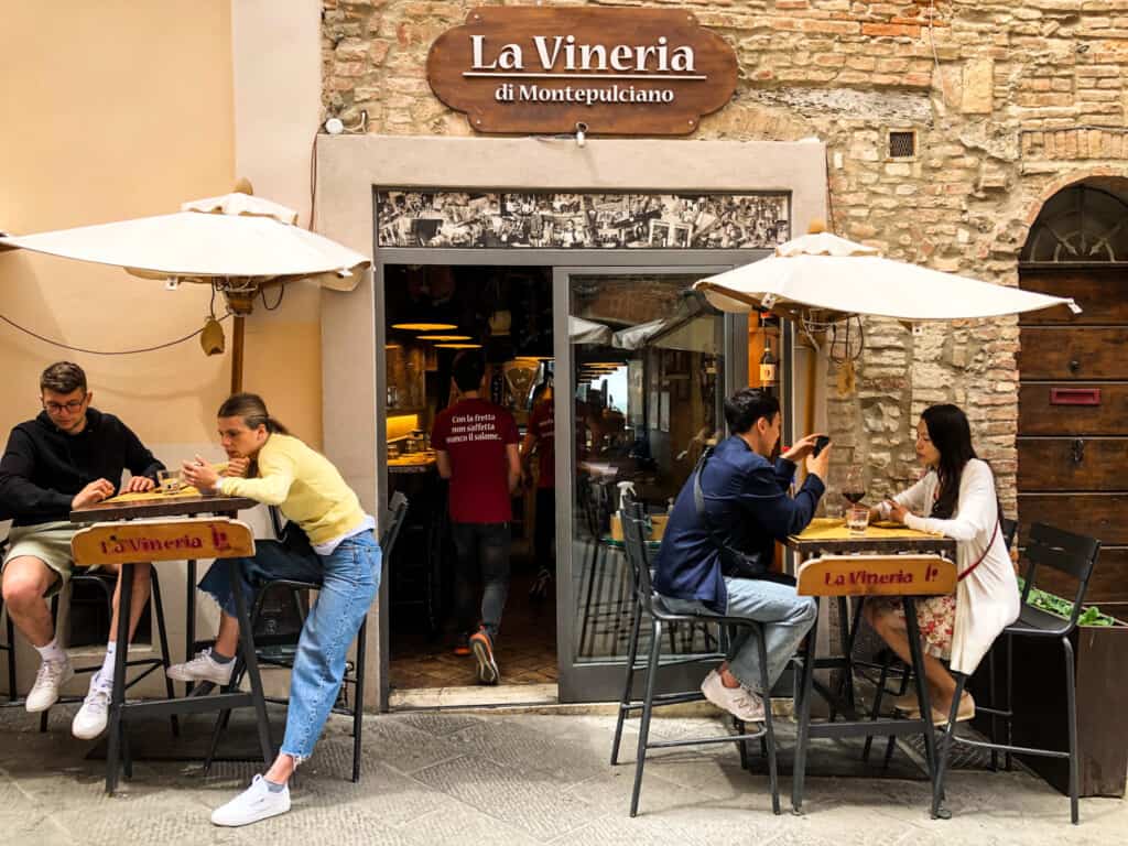 People sitting at small, tall tables with umbrellas in front of an enoteca in a small Italian town.