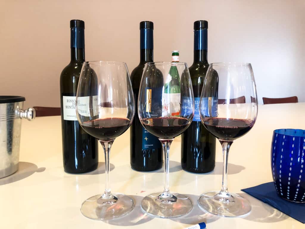 Three wine glasses with small pours of red wine on a white table. Behind the glasses are three bottles of red wine. One the right is a blue glass.