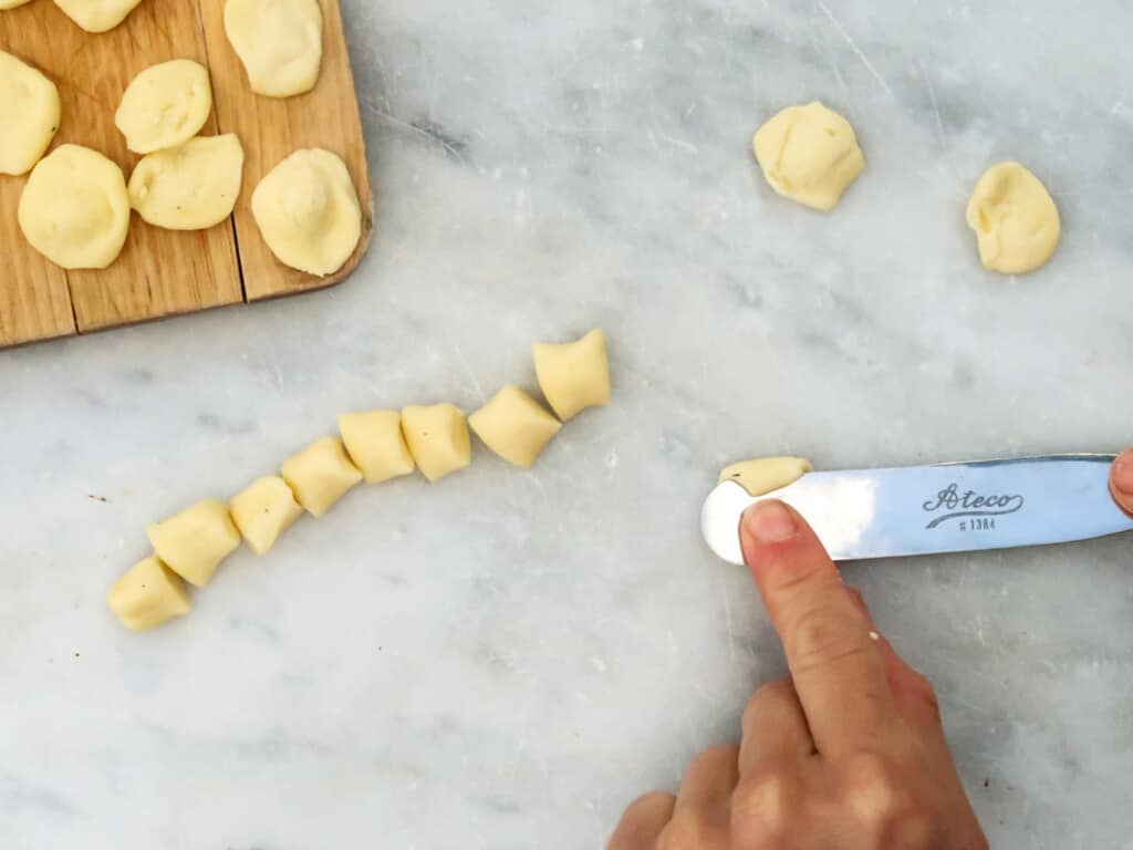 Finger pointing to orecchiette pasta being formed at the end of a metal spatula. Some dough and finished orecchiette sit on the table.