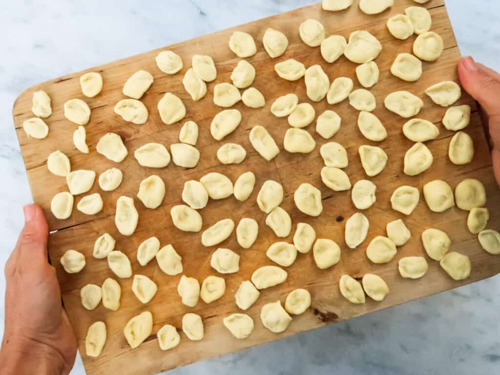 Hands holding wooden cutting board with orecchiette pasta drying.