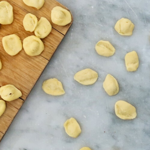 Part of wooden cutting board on left and white marble surface on right. Both places scattered with fresh orecchiette pasta.