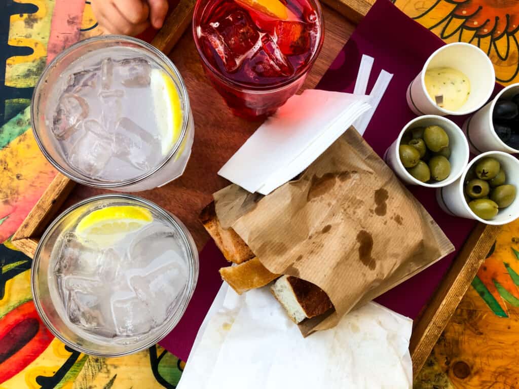 top view of a table with three soft drinks served on a tray with various small cups filled with snacks and a sack of bread.