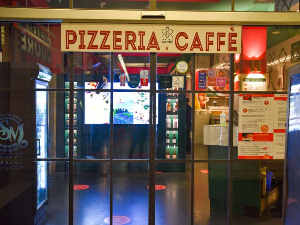 view of glass door with large sign in red written pizzeria fratelli cuore caffe with tv inside and ordering counter behind large glass doors.