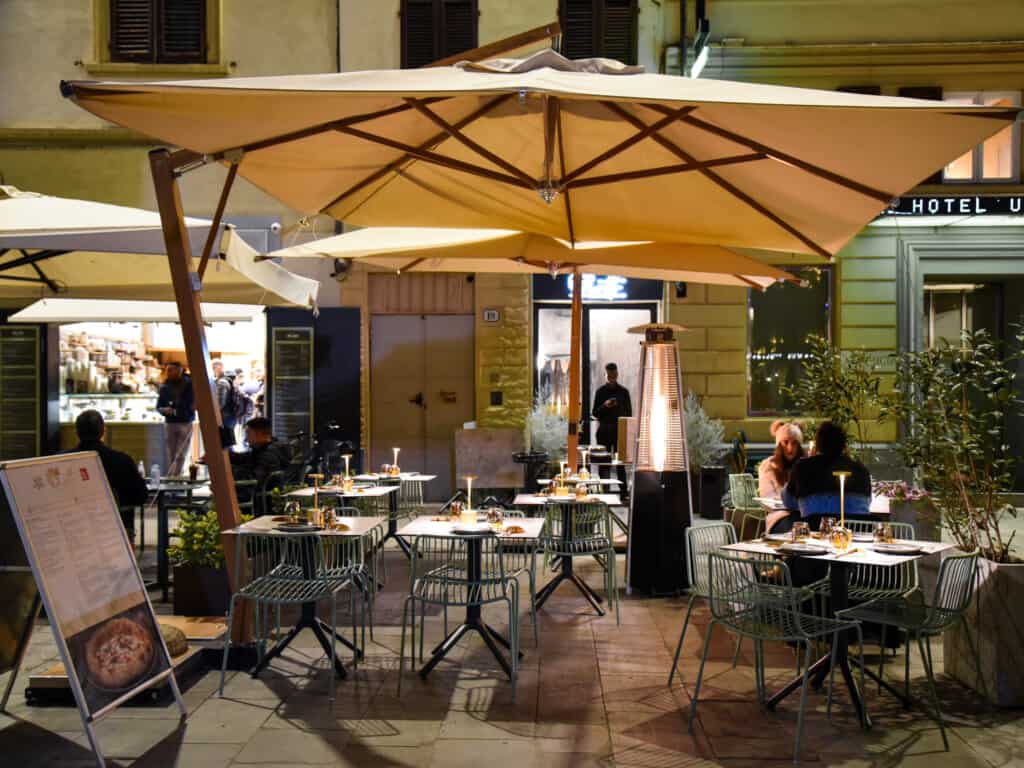 side view of pizzeria with large umbrella covering tables outdoors with a couple sitting at a table on the right hand side at dusk. 