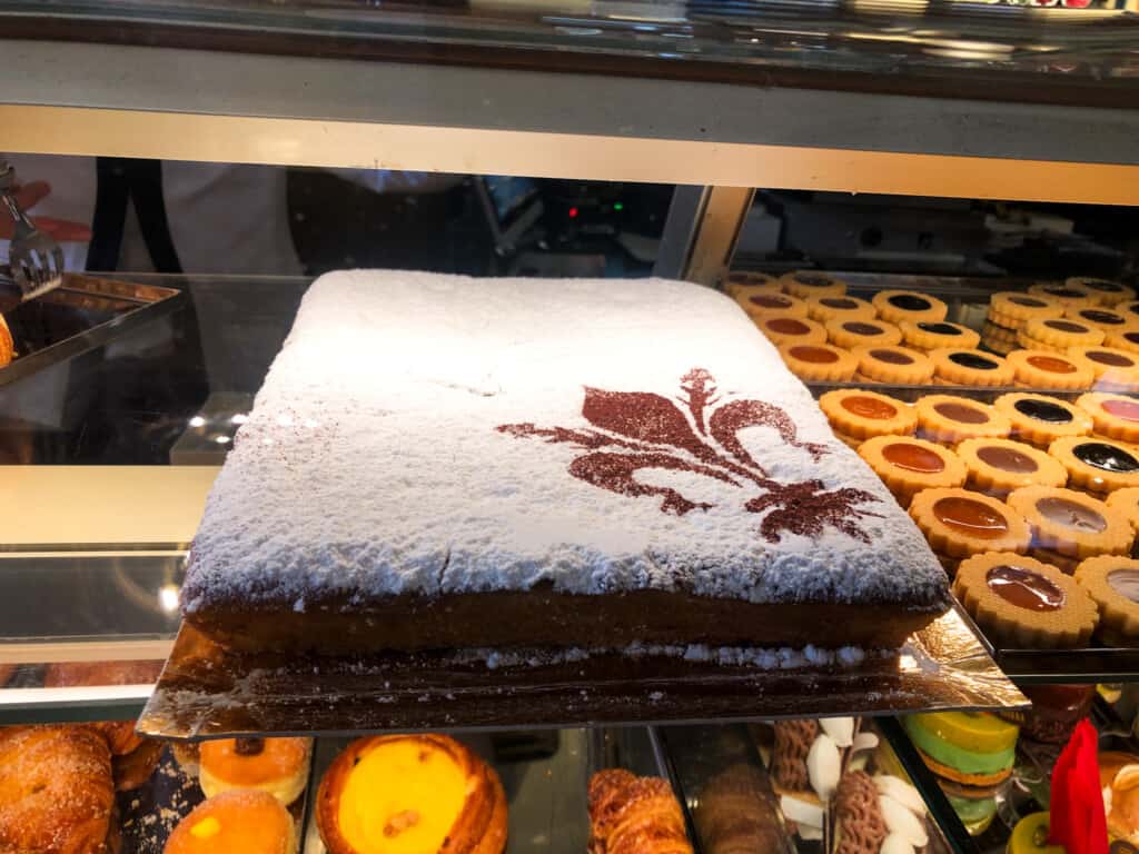 side view of large white sheet cake schiacciata alla fiorentina displayed in a glass case with a brown giglio flower in cocoa powder on right hand bottom corner of cake. 