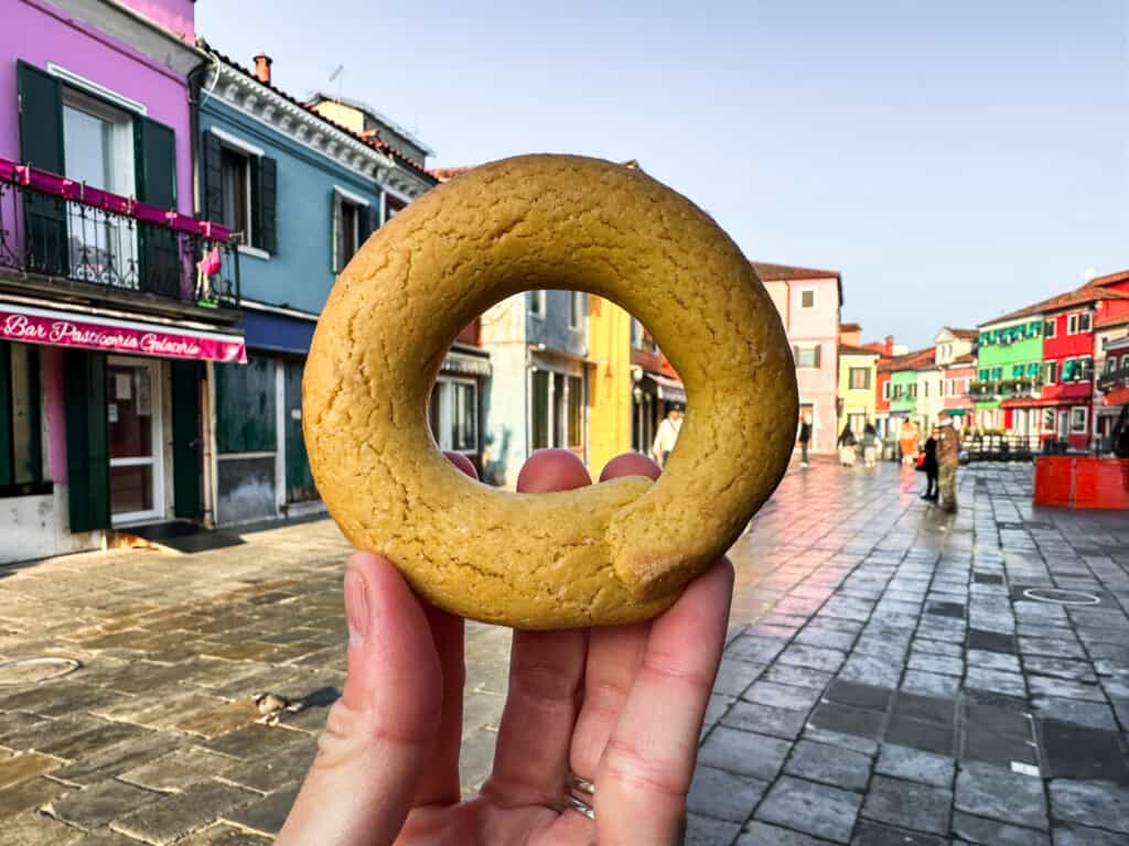 Hand holds up bussola cookie in front of piazza on Burano Island in Venice, Italy.