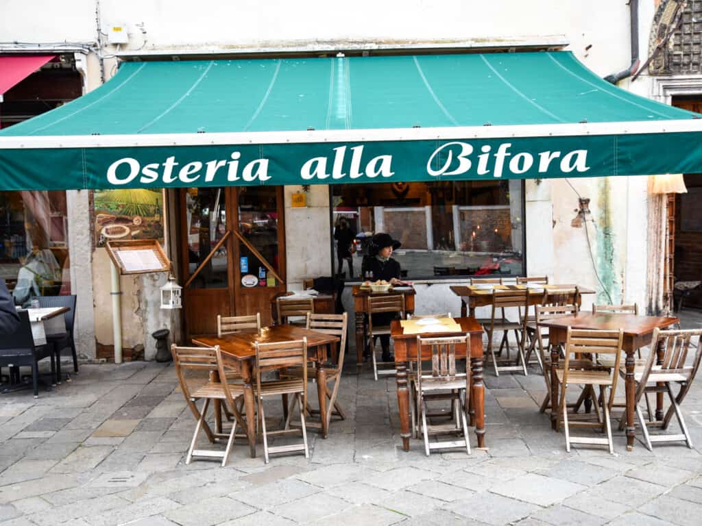 Green awning hangs above Osteria alla Bifora in Venice, Italy. Wooden tables and chairs out front with one woman eating.