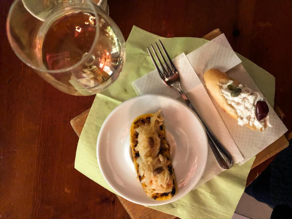 Plate with cicchetto plus another to the right on a napkin. Glass of white wine and fork. All are on wooden table.