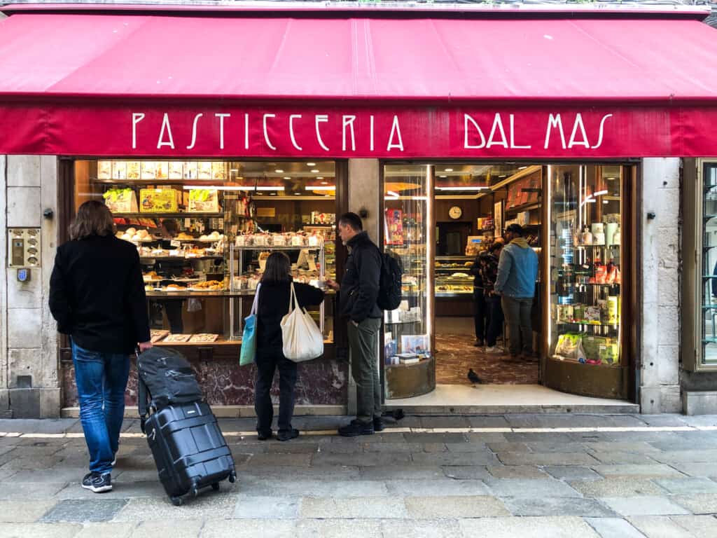 Red awning covers Pasticceria Dal Mais entry. Glass windows display pastries. Customers look in window and are inside in line.