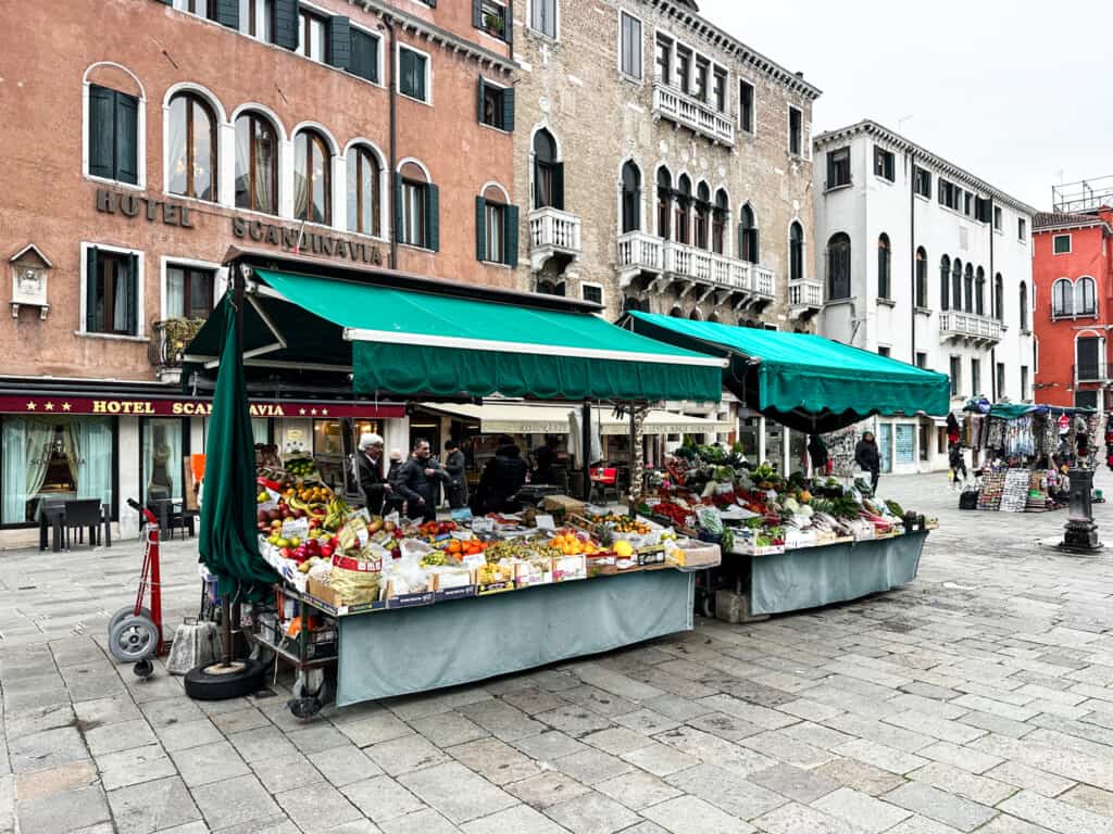 side view of large piazza in venice with a market stand with a green tent selling various fruits and vegetables outdoors in middle.