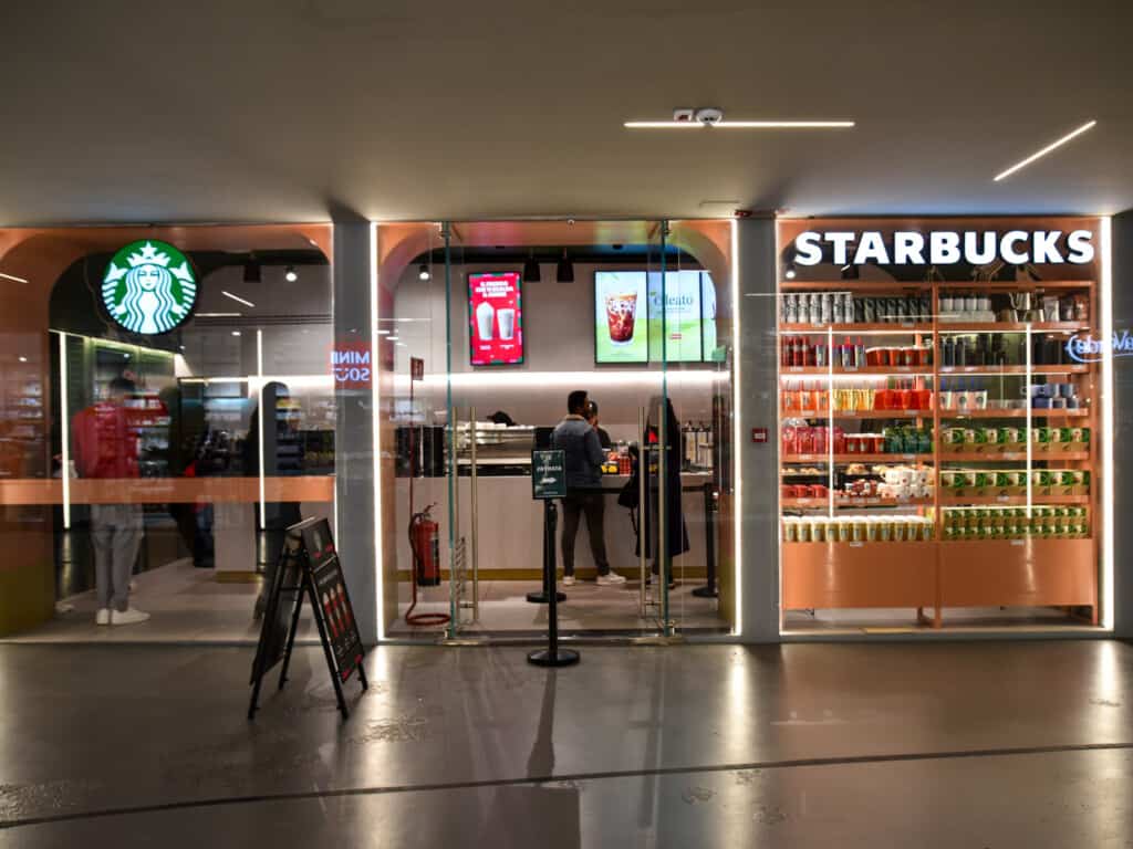 Entrance to Starbucks at the Florence train station in Italy. There are a few people in the shop ordering. Products in window display like mugs and cups.