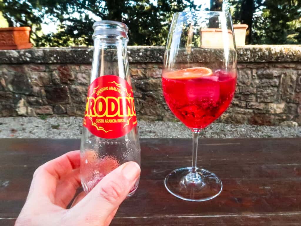 Hand holding empty small bottle of Crodino next to wine glass with crodino, ice and orange slice inside.