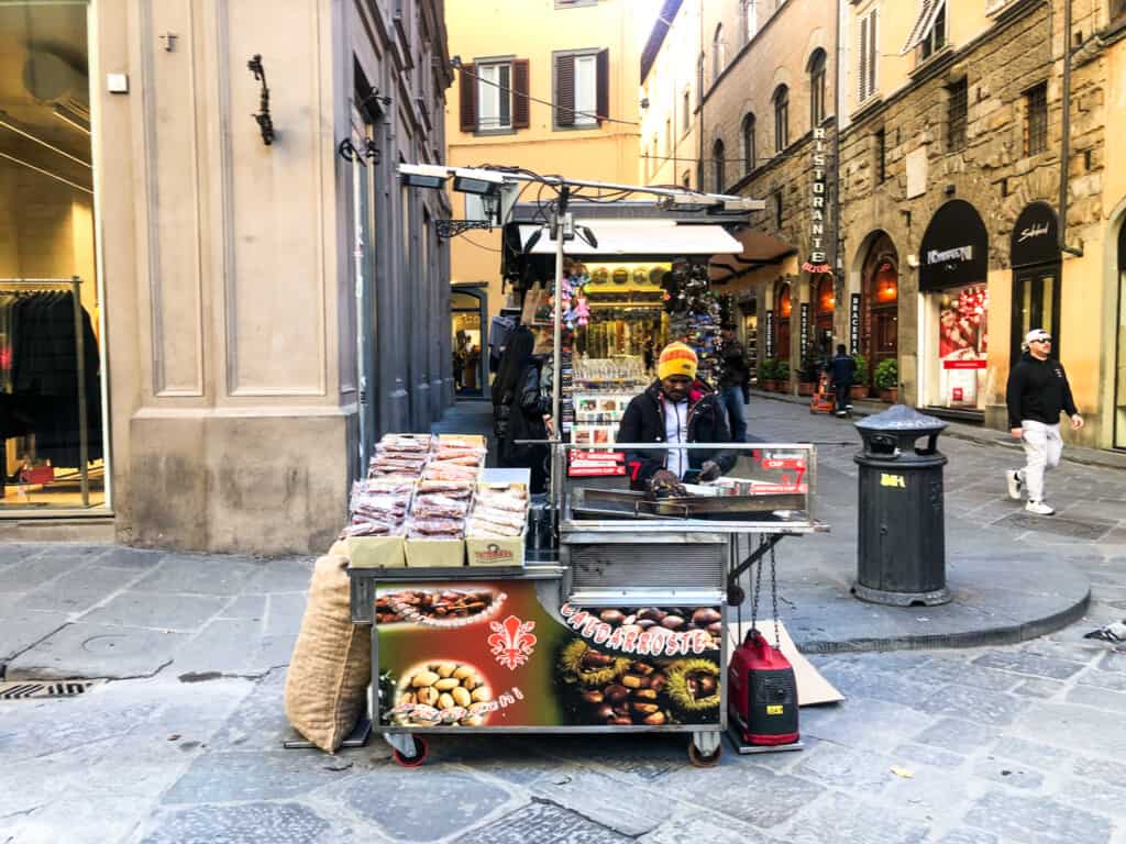 side view of man selling chestnuts on street in florence with yellow and brown buildings in background.