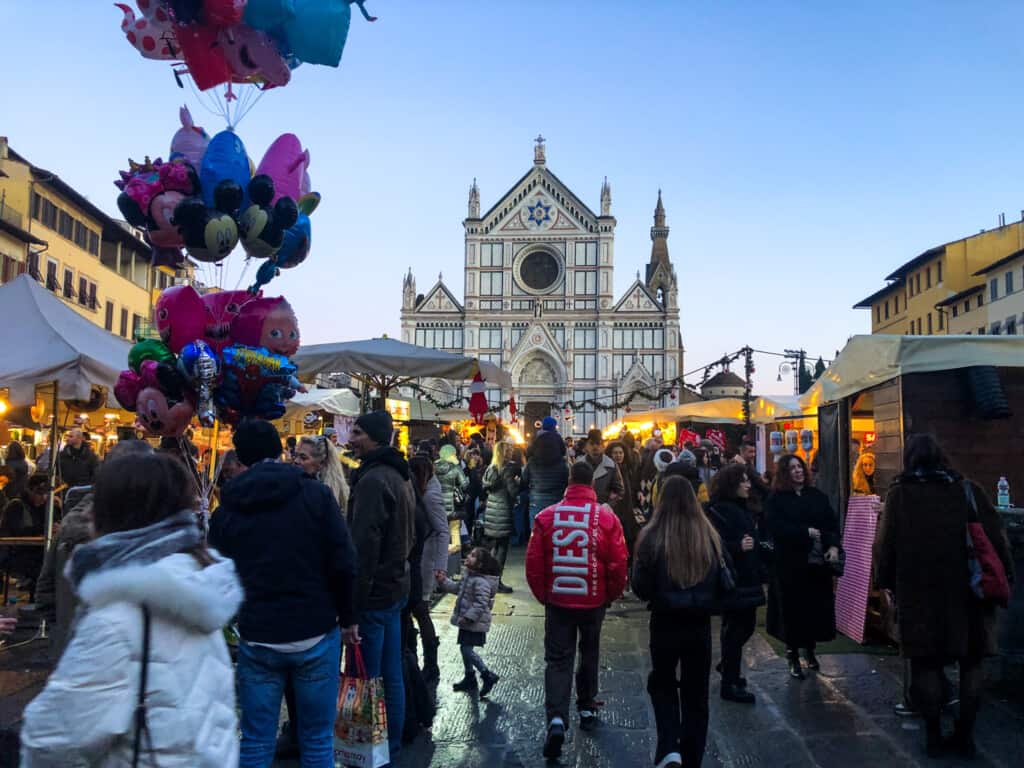 People walking around the Christmas market in Piazza Santa Croce in Florence, Italy. Balloons on left, church in background, market stalls lit up as evening arrives.