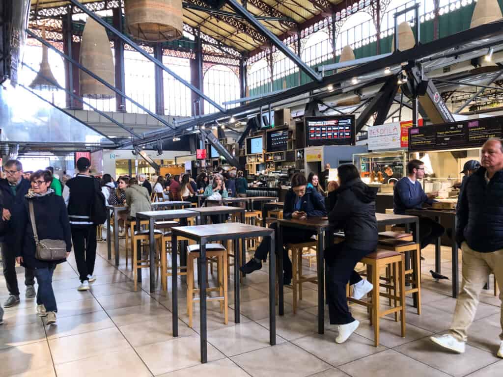 People wandering around market inside building with large windows and high ceilings. There are fixed food stalls with Italian food and wine. Some people sit at high tables with barstools.