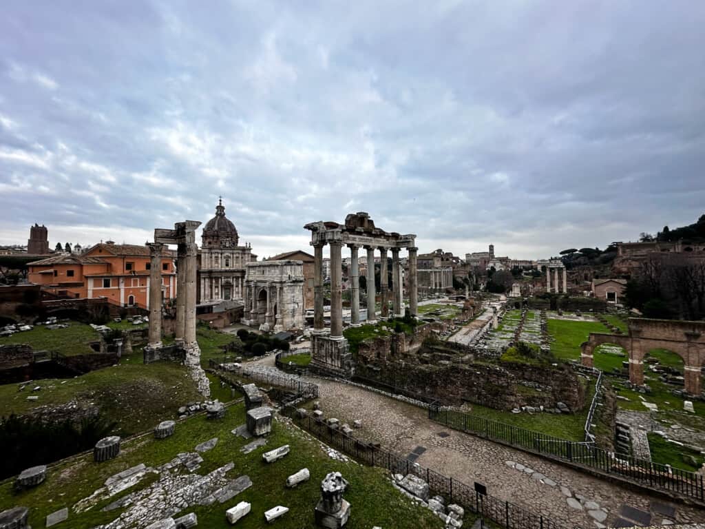 wide angle view of old roman archeological site in rome with tall structures in middle with grass and stones around them. 
