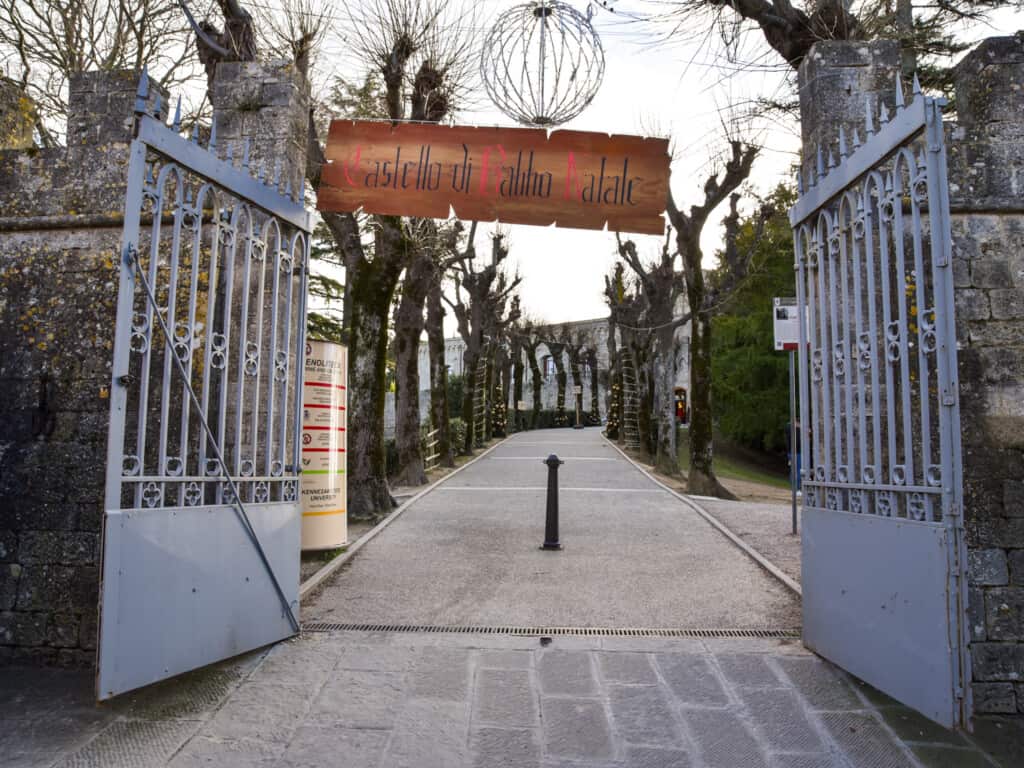 Wooden sign labeled 'Castello di Babbo Natale' hangs above open gate and tree-lined path in Montepulciano, Italy.