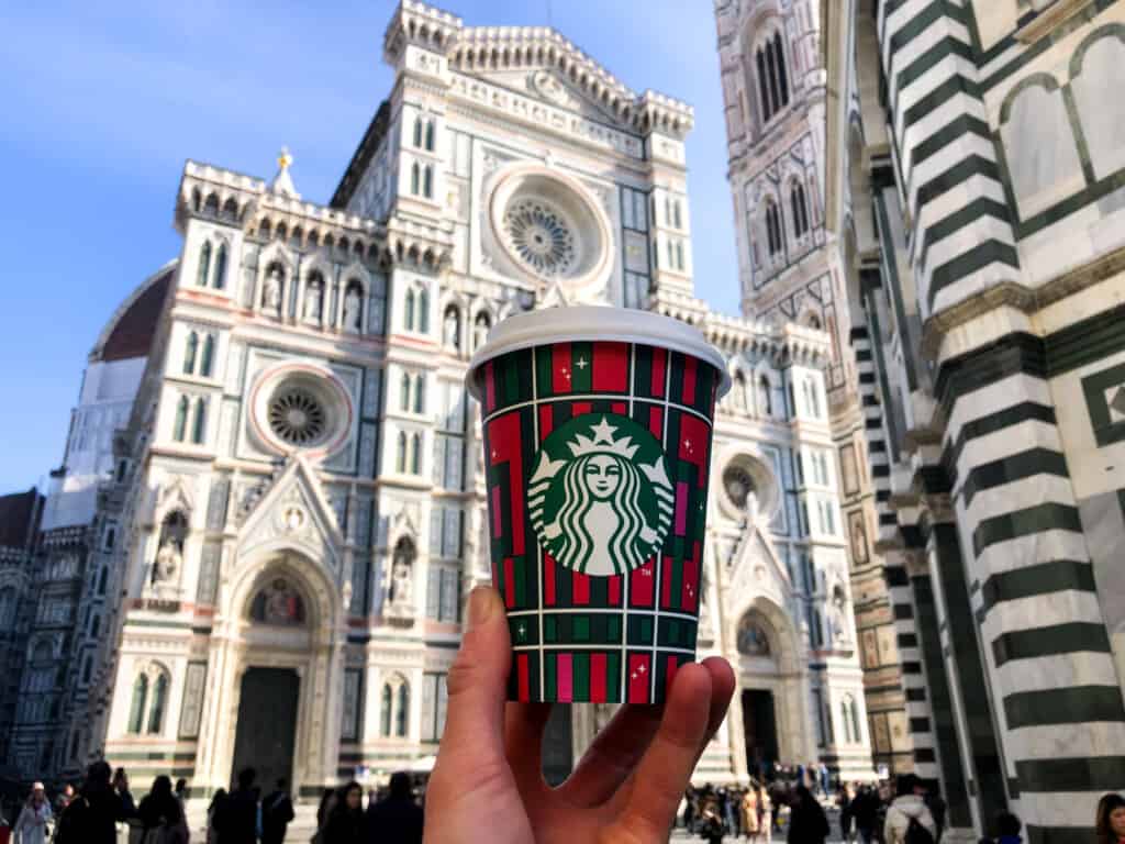 hand holding starbucks cup in front of Duomo in florence on a sunny day with blue skies in background.
