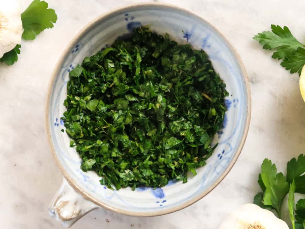 Bowl of gremolata surrounded by parsley leaves and garlic bulbs.