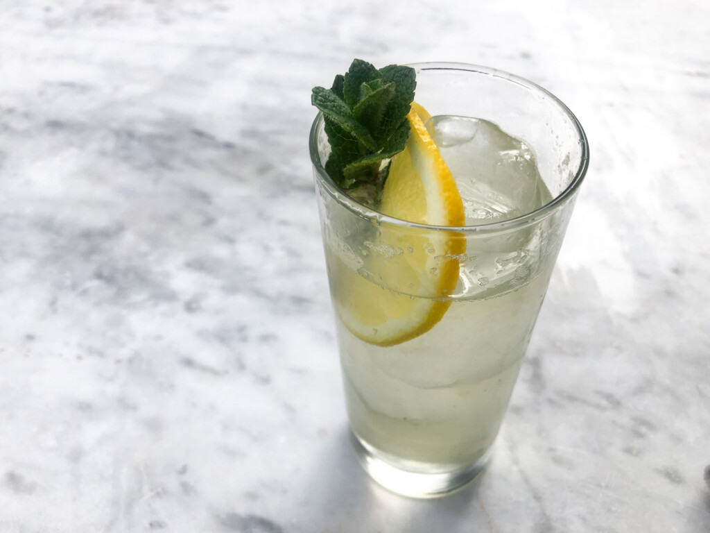 Glass with liquid, lemon slice, mint leaves, and ice on a white marble surface.